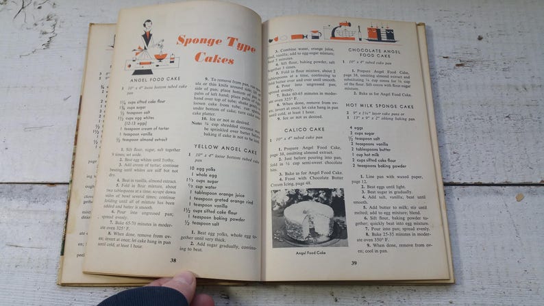 May include: Open vintage cookbook pages with recipes for sponge-type cakes. The pages are yellowed with age and feature illustrations and text. The book is open to pages 38 and 39, with a hand visible at the bottom.