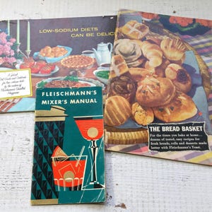 May include: A collection of vintage cookbooks with colorful covers. One book is titled "Fleischmann's Mixer's Manual" and another "The Bread Basket." The covers feature images of food and drinks, including baked goods and cocktails.
