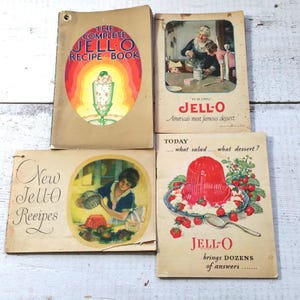 May include: Four vintage Jell-O recipe books with colorful covers. The covers feature illustrations of desserts, including a sundae, a bowl of Jell-O with strawberries, and a woman making Jell-O. The text on the covers includes "The Complete Jell-O Recipe Book", "It's so simple, Jell-O, America's most famous dessert", "Today... what salad... what dessert?", and "New Jell-O Recipes".
