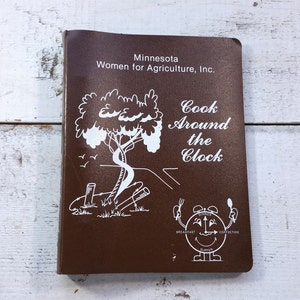 May include: A brown cookbook with the title "Cook Around the Clock" and a white illustration of a clock with a face and hands. The book is from the Minnesota Women for Agriculture, Inc.