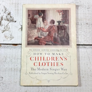 May include: A vintage book titled "How to Make Children's Clothes The Modern Singer Way" published by Singer Sewing Machine Co., Inc. The cover features an illustration of a woman helping a young girl put on a pair of shoes.