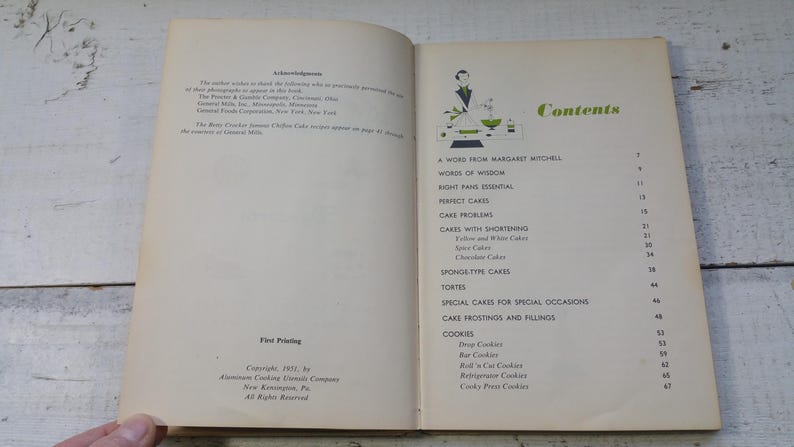 May include: Open vintage cookbook with a contents page featuring a green and white illustration. The table of contents lists cake recipes, including spice cakes and chocolate cakes, and cookie recipes.