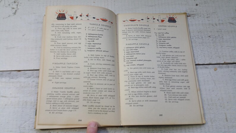 May include: An open vintage cookbook page featuring recipes for Vanilla Souffle, Pineapple Tapioca, Orange Souffle, Chocolate Souffle, and Coffee Souffle. The text is printed in black ink on off-white paper with small red and black illustrations.