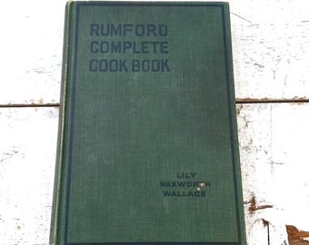 May include: A vintage cookbook titled "Rumford Complete Cook Book" with a dark green cover. The book's title is printed in white, with the author's name, Lily Haxworth Wallace, at the bottom. The book is on a white wooden surface.