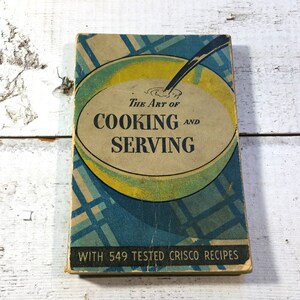 Puede incluir: Libro de cocina vintage con una ilustración azul y amarilla de un tazón y una cuchara en la portada. El título es "The Art of Cooking and Serving" con el texto "With 549 Tested Crisco Recipes".