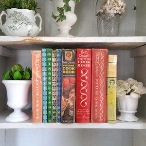 May include: A shelf displays a collection of vintage cookbooks, including several "Betty Crocker's Picture Cook Book" editions. The books are in various colors, with red, orange, and yellow spines. Decorative vases with plants and flowers are also on the shelf.