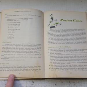 May include: Open vintage cookbook pages with text and illustrations. The left page lists baking measurements, while the right page features the title "Perfect Cakes" with a green and white illustration. The book is open on a white wooden surface.