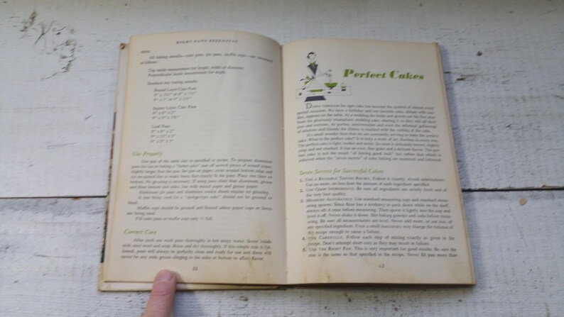 May include: Open vintage cookbook pages with text and illustrations. The left page lists baking measurements, while the right page features the title "Perfect Cakes" with a green and white illustration. The book is open on a white wooden surface.