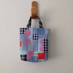 Könnte beinhalten: Eine Patchwork-Tragetasche mit verschiedenen blauen Stoffen, darunter Streifen, Karos und Unifarben, hängt an einem Holzhaken. Die Tasche hat rosa und rote geometrische Formen. Die Griffe sind ein schwarz-weißes Karomuster.