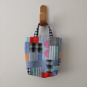 May include: A patchwork tote bag with a variety of blue, red, and pink fabric squares. The bag has a gingham pattern on the handles and is hanging on a wooden hook. The bag is made of various fabrics.
