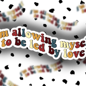 May include: A sticker with the text "I'm allowing myself to be led by love" in a retro font with colorful letters. The sticker is on a white background with black dots.