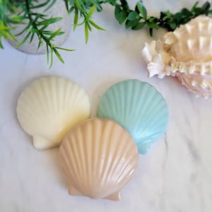 May include: Three decorative seashell-shaped soaps in white, light brown, and light blue. The soaps are arranged on a white marble surface.