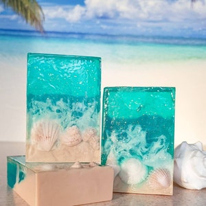 K&ouml;nnte beinhalten: Drei rechteckige Seifenst&uuml;cke mit Strandthema. Die Seife ist klarblau mit wei&szlig;en Wirbeln und eingebetteten Muscheln. Die untere Seife ist hellbeige.