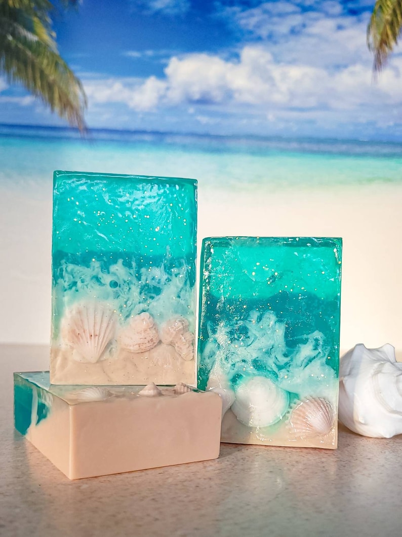 K&ouml;nnte beinhalten: Drei rechteckige Seifenst&uuml;cke mit Strandthema. Die Seife ist klarblau mit wei&szlig;en Wirbeln und eingebetteten Muscheln. Die untere Seife ist hellbeige.