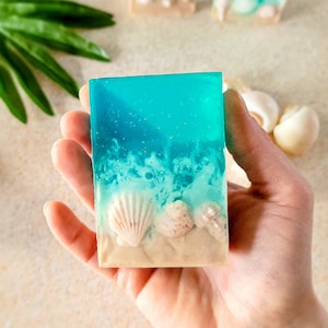 K&ouml;nnte beinhalten: Eine blaue und wei&szlig;e Seife mit einer Strandszene und Muscheln. Die Seife liegt in einer Hand, bereit zum Gebrauch.