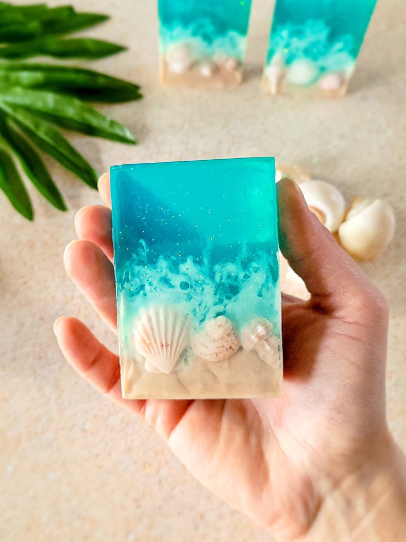 K&ouml;nnte beinhalten: Eine blaue und wei&szlig;e Seife mit einer Strandszene und Muscheln. Die Seife liegt in einer Hand, bereit zum Gebrauch.