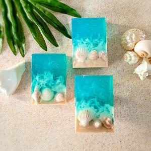 K&ouml;nnte beinhalten: Drei rechteckige Seifenst&uuml;cke mit einem blauen und wei&szlig;en Ozeandesign und wei&szlig;en Muscheln, die in die Seife eingebettet sind. Die Seife liegt auf einer hellbeigen Oberfl&auml;che.