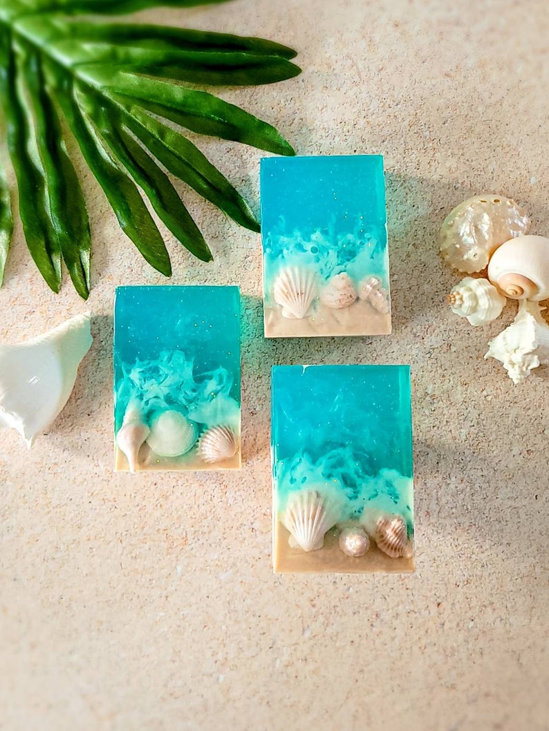 K&ouml;nnte beinhalten: Drei rechteckige Seifenst&uuml;cke mit einem blauen und wei&szlig;en Ozeandesign und wei&szlig;en Muscheln, die in die Seife eingebettet sind. Die Seife liegt auf einer hellbeigen Oberfl&auml;che.