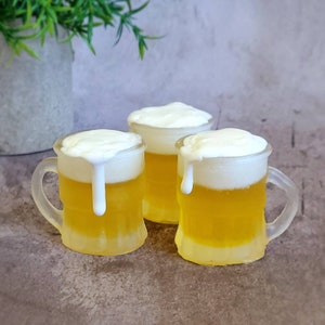 May include: Three miniature glass beer mugs with white foam on top. The mugs are filled with a yellow liquid that resembles beer.