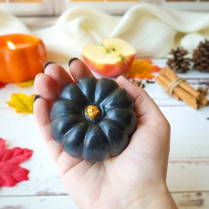 May include: A black pumpkin-shaped soap with a gold accent in the center. The soap is being held in a person's hand.