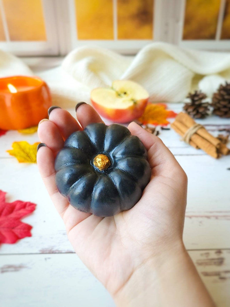 May include: A black pumpkin-shaped soap with a gold accent in the center. The soap is being held in a person's hand.