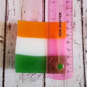 May include: A rectangular bar of soap with three layers of colour: orange, white, and green. The soap is measured against a ruler, showing it is approximately 7 cm long.