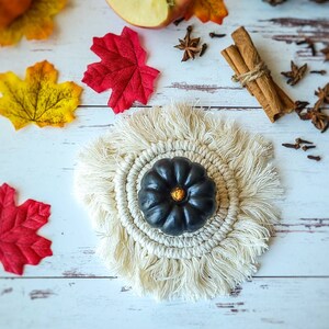 May include: A black pumpkin with a gold stem sits on a white crocheted coaster with fringe. The coaster is on a white wooden surface with fall decor, including red and yellow leaves, cinnamon sticks, and star anise.