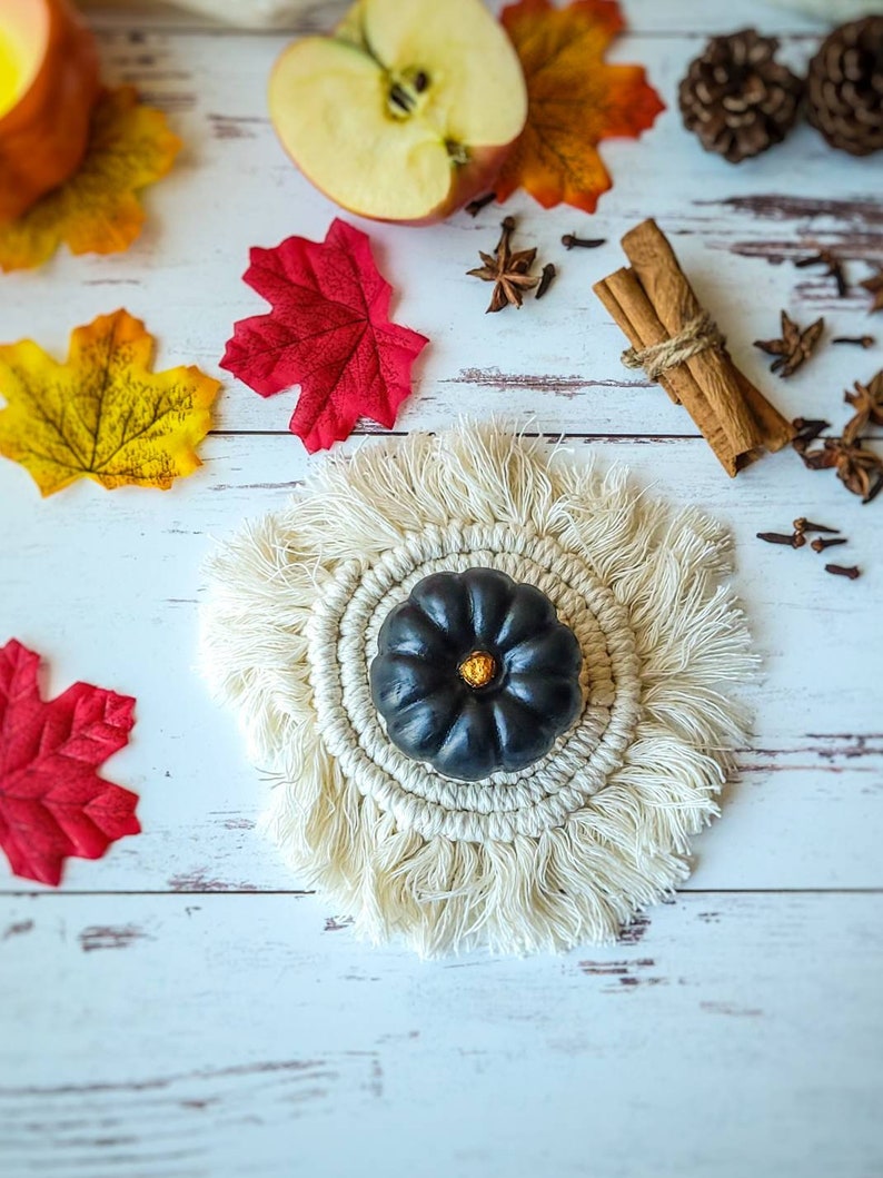 May include: A black pumpkin with a gold stem sits on a white crocheted coaster with fringe. The coaster is on a white wooden surface with fall decor, including red and yellow leaves, cinnamon sticks, and star anise.