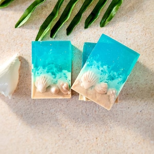 K&ouml;nnte beinhalten: Drei Seifenst&uuml;cke mit Strandmotiv. Die Seife ist blau und wei&szlig; mit einer braunen Schicht am Boden. Die Seife hat Muscheln eingebettet.