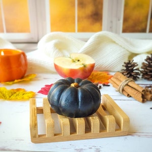 May include: A black pumpkin-shaped soap bar sits on a wooden soap dish. The soap dish is on a white wooden surface. There is a half-eaten apple, cinnamon sticks, and fall leaves in the background.