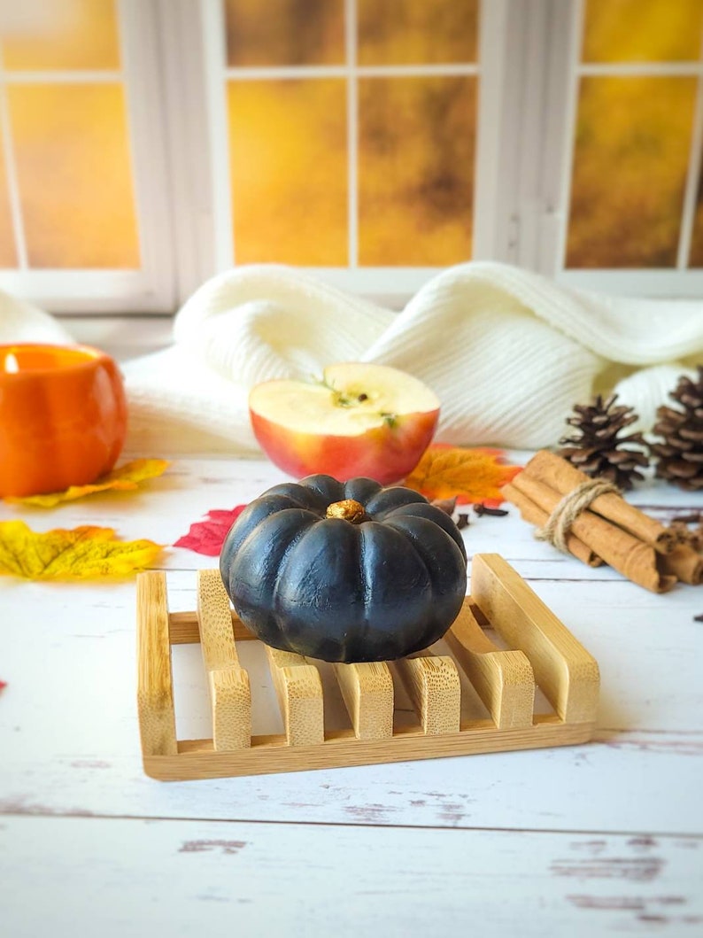 May include: A black pumpkin-shaped soap bar sits on a wooden soap dish. The soap dish is on a white wooden surface. There is a half-eaten apple, cinnamon sticks, and fall leaves in the background.