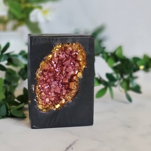 May include: A black soap bar with a pink and gold geode design. The geode is a cluster of crystals with a shiny, metallic finish.