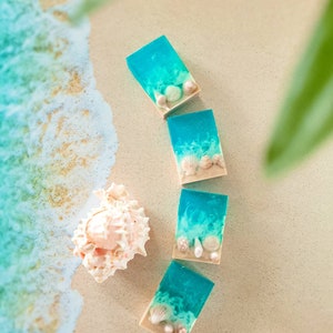 K&ouml;nnte beinhalten: Vier t&uuml;rkisblaue Seifenst&uuml;cke mit eingebetteten wei&szlig;en und braunen Muscheln. Die Seifenst&uuml;cke sind in einer Reihe auf einer hellbraunen Oberfl&auml;che angeordnet.