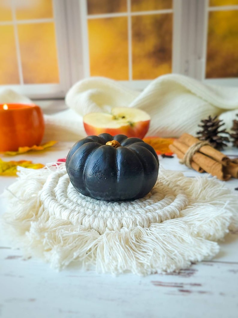 May include: A black pumpkin with a gold stem sits on a white crocheted coaster with fringe. The coaster is on a white wooden surface. There is a red apple and cinnamon sticks in the background.