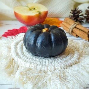May include: A black pumpkin with a gold stem sits on a white crocheted coaster with fringe. The pumpkin is on a white wooden surface with a red and orange leaf in the background.