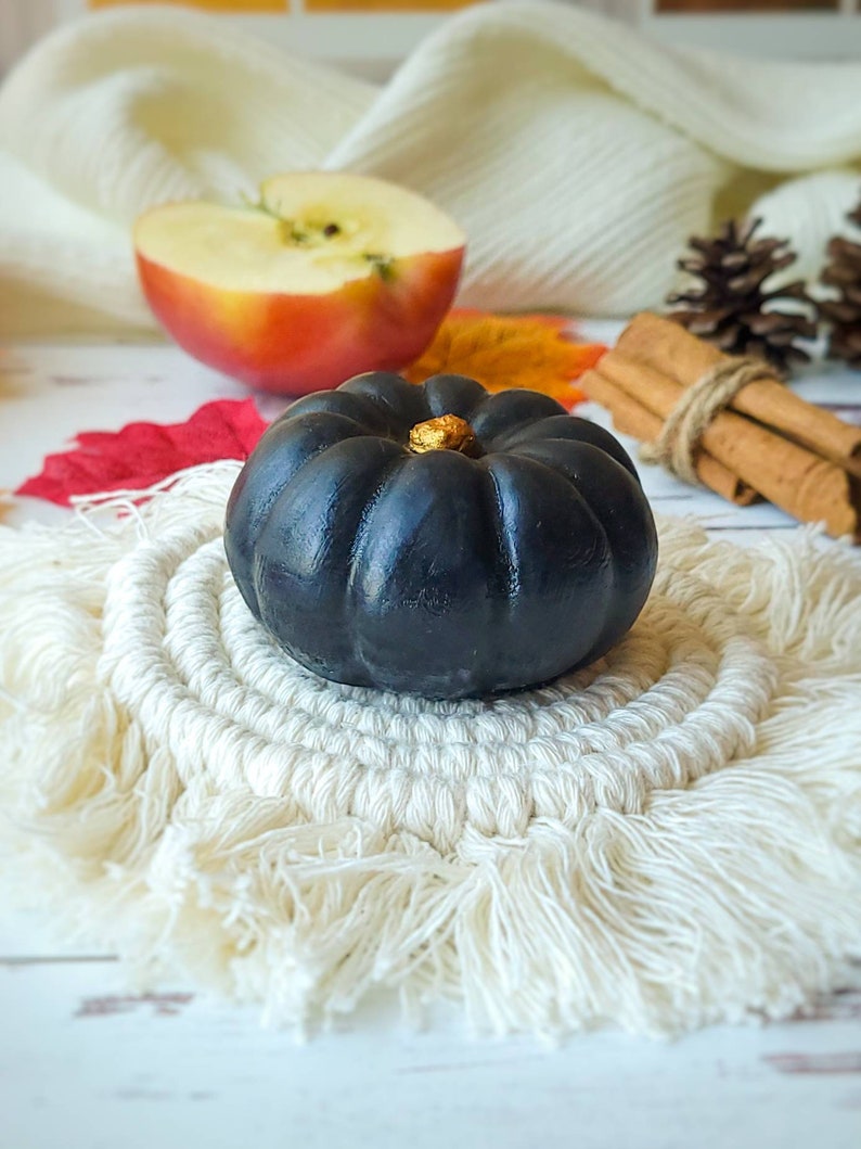 May include: A black pumpkin with a gold stem sits on a white crocheted coaster with fringe. The pumpkin is on a white wooden surface with a red and orange leaf in the background.