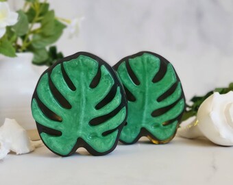 May include: Two decorative cabinet knobs in the shape of a monstera leaf. The knobs are green with black outlines and a gold-colored edge. The knobs are set against a white background with greenery and seashells.