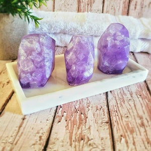 May include: Three purple amethyst-shaped soap bars on a white marble soap dish. The soap bars have a glittery finish.