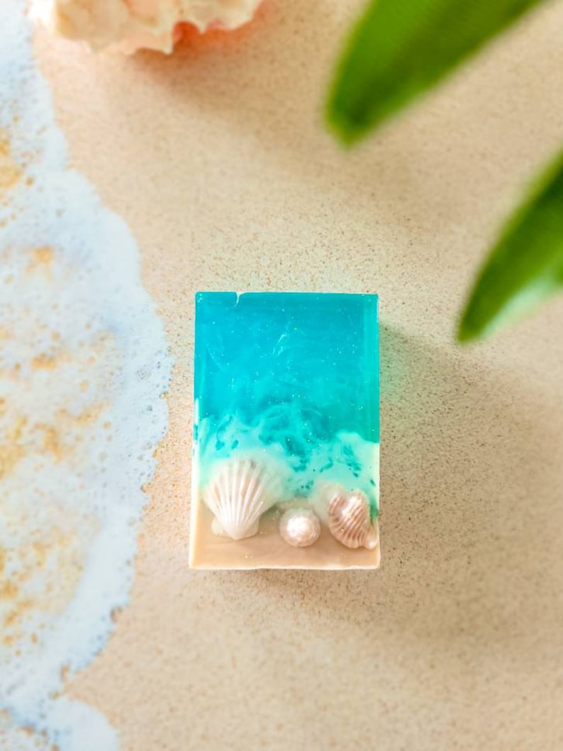 K&ouml;nnte beinhalten: Eine rechteckige Seife mit einem blauen und wei&szlig;en Design, das an eine Strandszene erinnert. Die Seife zeigt wei&szlig;e Muscheln und eine Perle.