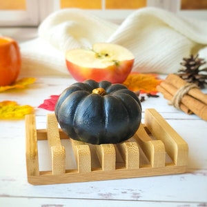 May include: A black pumpkin-shaped soap bar sits on a wooden soap dish. The soap dish is on a white surface with fall-themed decorations, including a red apple, cinnamon sticks, and pine cones.