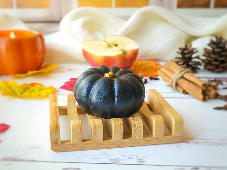 May include: A black pumpkin-shaped soap bar sits on a wooden soap dish. The soap dish is on a white surface with fall-themed decorations, including a red apple, cinnamon sticks, and pine cones.