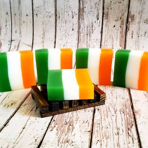 May include: Five bars of soap with green, white and orange stripes. The bars are arranged in a row on a wooden surface. One bar is on a small wooden stand.