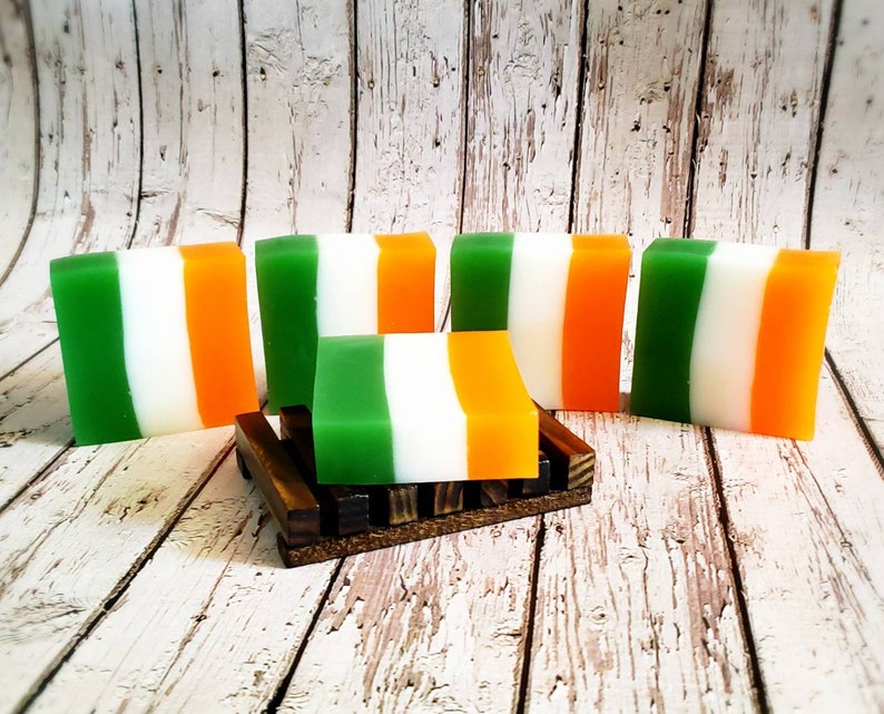 May include: Five bars of soap with green, white and orange stripes. The bars are arranged in a row on a wooden surface. One bar is on a small wooden stand.