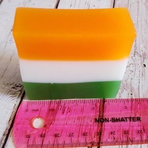 May include: A rectangular bar of soap with three layers of colour: orange, white, and green. The soap is measured against a pink ruler, showing it is approximately 5 cm long.