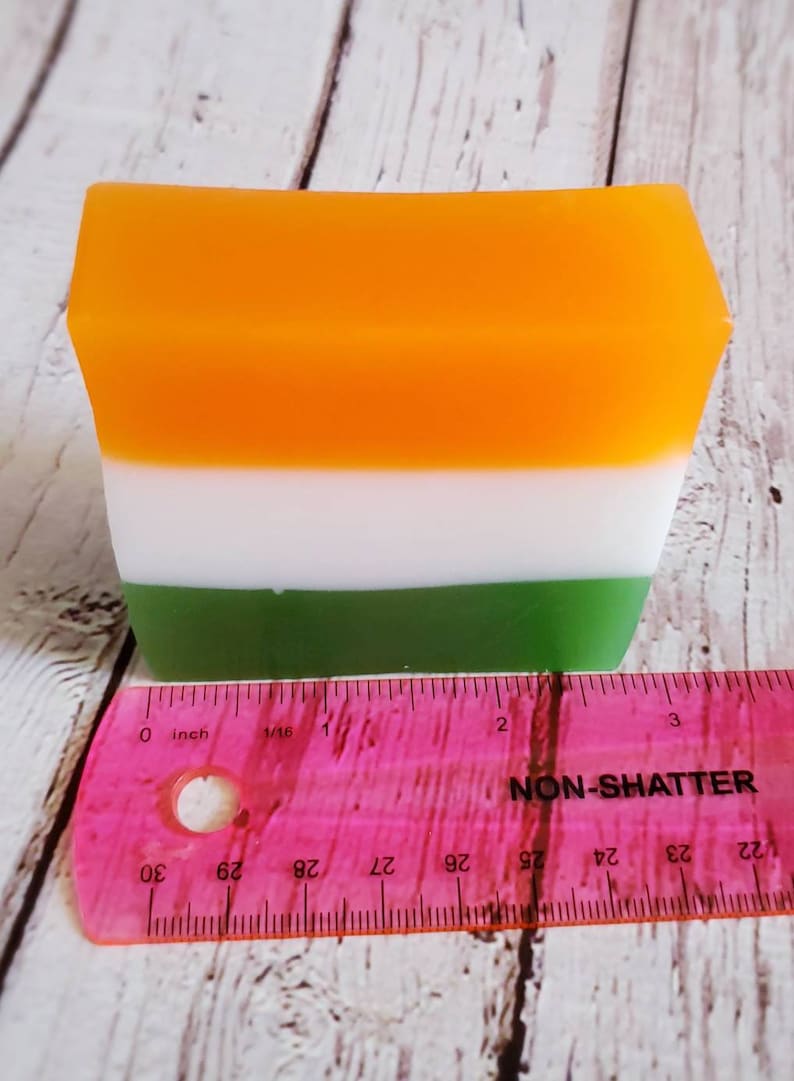 May include: A rectangular bar of soap with three layers of colour: orange, white, and green. The soap is measured against a pink ruler, showing it is approximately 5 cm long.