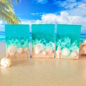 K&ouml;nnte beinhalten: Drei St&uuml;ck t&uuml;rkisblaue Seife mit wei&szlig;en Wirbeln und Muscheln, die in die Seife eingebettet sind. Die Seife liegt auf einer sandigen Oberfl&auml;che.