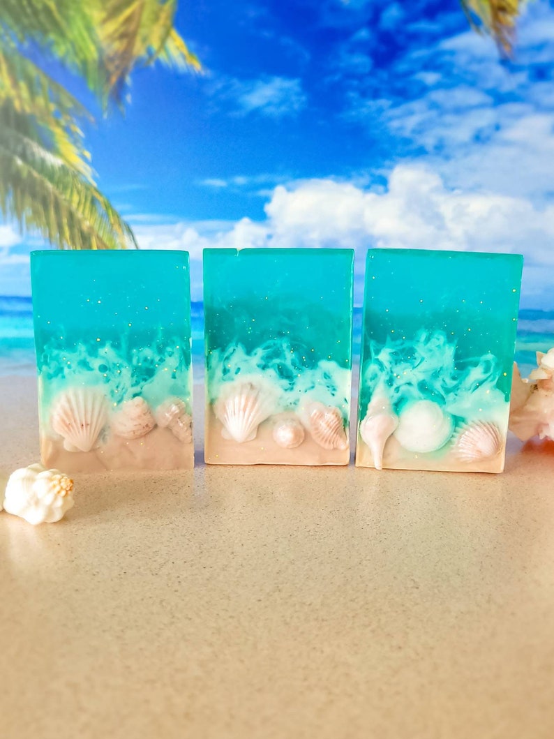 K&ouml;nnte beinhalten: Drei St&uuml;ck t&uuml;rkisblaue Seife mit wei&szlig;en Wirbeln und Muscheln, die in die Seife eingebettet sind. Die Seife liegt auf einer sandigen Oberfl&auml;che.