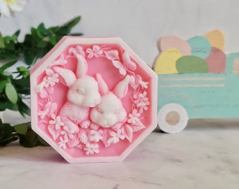 May include: A pink octagonal soap with a white border features a detailed design of two white rabbits nestled in a wreath of pink flowers and leaves.