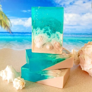 K&ouml;nnte beinhalten: Drei handgemachte Seifen mit Strandthema. Die obere Seife ist klarblau mit wei&szlig;en Wirbeln und eingebetteten Muscheln. Die beiden unteren Seifen sind hellbraun mit einem klaren blauen Wirbel. Die Seife liegt auf einem Sandstrand mit einer Muschel im Hintergrund.