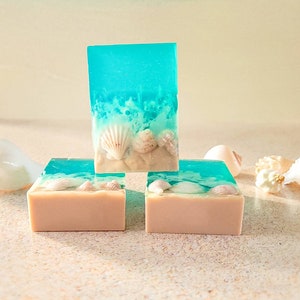 K&ouml;nnte beinhalten: Drei Seifenst&uuml;cke mit Strandmotiv. Die obere Seife ist blau und gr&uuml;n mit wei&szlig;en Muscheln, die in die Seife eingebettet sind. Die beiden unteren Seifen sind beige mit wei&szlig;en Muscheln, die in die Seife eingebettet sind.